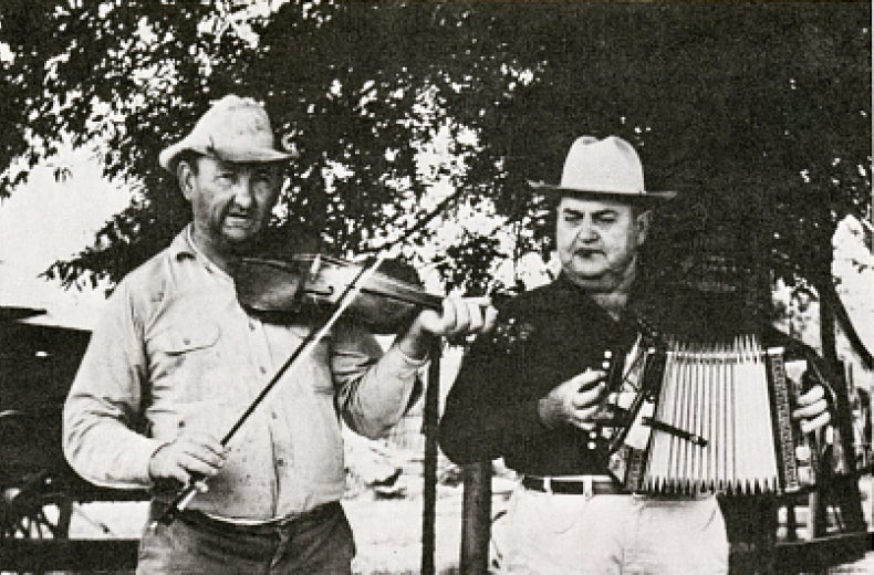 Two men standing in the foreground. The left person is playing a violin while the right person is playing an accordion. 