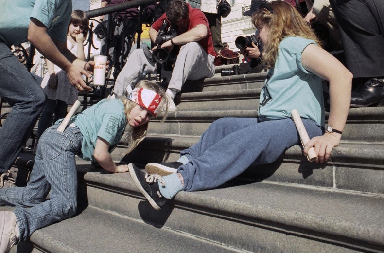 Jennifer Keelan-Chaffins wears a bandana as she crawls up the U.S. Capitol steps on her hands and knees. On her left is a man bending over and on her right a woman moves up the stairs on her back. All three protesters wear matching light blue shirts from the ADAPT organization. In front of them are media personnel with cameras and microphones recording the protest. ​​​​​[Photo Credit: Associated Press]