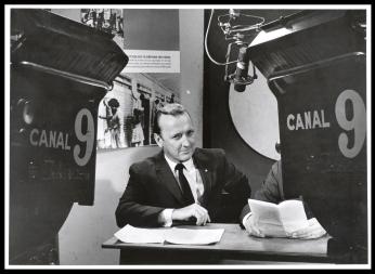 Grant sits at a desk in a newsroom. He is sharply dressed with a suit and tie. He looks intently at someone or something not captured in the picture. 