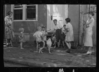 A black-and-white photo of young children, two women, and a dog standing around a stoop