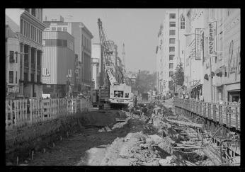 A black-and-white photo of Metro construction. There is a hole in the middle of a street with buildings on either side. There are construction workers and vehicles in and around the hole.