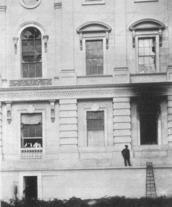 Exterior view of the west wall of the Capitol. Windows on the top floor have shattered glass, and one on the main floor is completely blown out and stained black from smoke.
