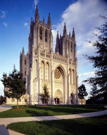 The front of the National Cathedral, with its two front towers and spires tall against a blue sky. The front of the building is lit by the sun.