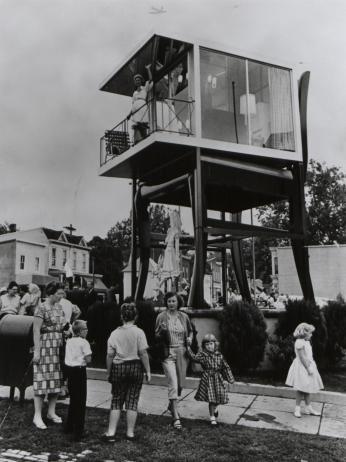A black-and-white photo of a giant chair with a glass house on top of it, people are gathered at the base of the chair looking up.