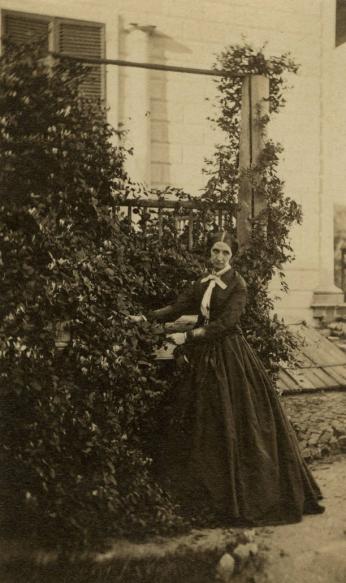 A photo of Sarah Tracy standing under a climbing plant in front of one of Mount Vernon's outbuildings
