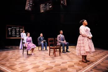 A scene from Tempestuous Elements. Several actors playing students are seated in chairs in the middle of the stage. Dr. Cooper watches from behind as one student stands and addresses the class.