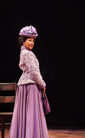 Gina Daniels as Anna Julia Cooper. She is standing in the spotlight on a dark stage wearing a purple skirt, jacket, and hat in the early 1900s style. 