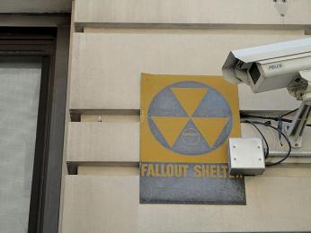 Photo of black and yellow "fallout shelter" sign on building facade, next to surveillance camera.
