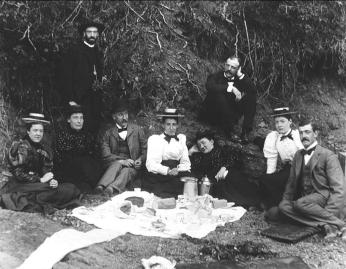 Black and white photo of well-dressed 19th-century men and women picnicking in a creek, looking at the camera