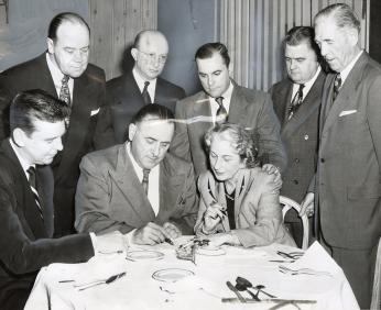 A group of people gather as Mrs. Florence Menendez signs the lease of Turner's Arena to Vince McMahon Sr., seated two spots from her right. Her husband, Gabe Menendez, stands over her.