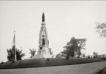 Black and white ground-level photo from 1900s of a cemetery with a concrete monument supporting statue of a man on top.