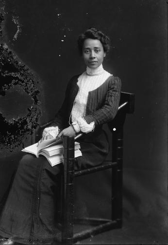 Educator-Writer Anna Julia Cooper photographed seated with book in hand and dressed in white collared black gown