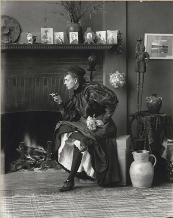 Full body photograph of Frances Benjamin Johnston seated, wearing a dark formal dress and holding a cigarette, 1896. (Source: Library of Congress)