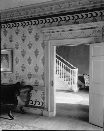 A viewpoint from the inside of a home, depicting a staircase through a doorway, in the foreground a room containing a daybed. 