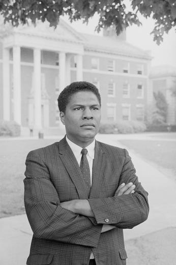 Professor Nathan Hare posing in front of Howard University's Douglass Hall dressed in suit with arms crossed.