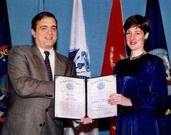 Man and woman posing and smiling for camera in front of flags while woman holds open certificate.