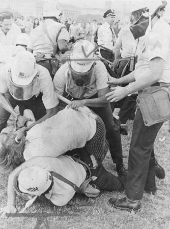 A group of police arrest a Yippie protestor, pushing him to the ground on the National Mall during Honor America Day celebrations.