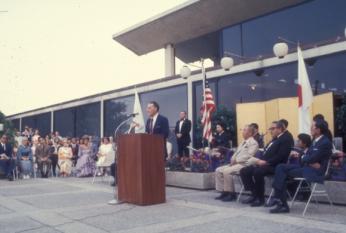 Photo of American man speaking at podium in front of building with American and Japanese flags, and American and Japanese politicians seated behind him, including Secretary of State Henry Kissinger.