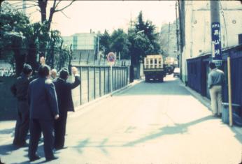Photo of group of Japanese men in business attire waving goodbye to a truck carrying a crate down a road.
