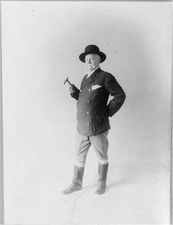 Black and white portrait of Edward Payson Weston looking at the camera holding a cane and wearing a double breasted suit jacket, mid-calf length boots, and a wide brim hat.