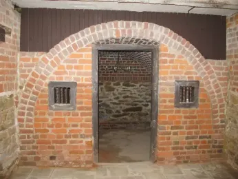 A Photograph of a brick arched entry way with a wooden frame in the middle. Behind the open door is a empty room with a small chandelier hanging from the ceiling