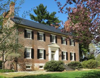 The picture is of a 2 story brick house with a black roof and 4 chimneys, with two on either side. The front of the house is slightly obscured by large bushes that sit along the front and side, and trees which are scattered around the house