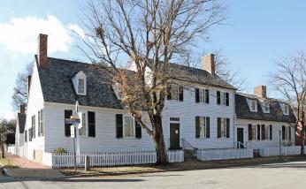 A photo of a large white house with black roof and shutters. The center of the house has a second floor and on either side of it dormer windows are unevenly placed along the roof