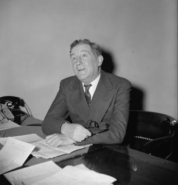 Black and white photo of man sitting at desk covered with papers and looking up toward the ceiling.