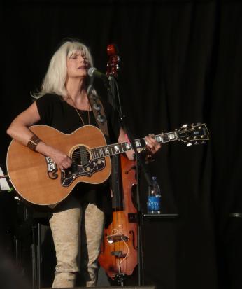 EmmyLou Harris standing, holding a guitar and singing into a microphone.
