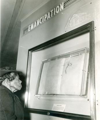 Elderly African American woman look at document in case on the wall. The word Emancipation is displayed over the case.