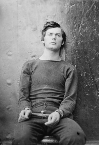 Black and white photo of young man sitting against a wall and wearing handcuffs.