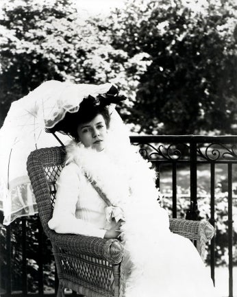 Black and white photo of young woman in white dress and white parasol sitting in chair.