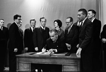 President Lyndon Johnson signs bill as a group of onlookers encircle his desk.