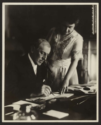 Black and white photo of Edith standing over Woodrow Wilson and working on documents. Source: Encyclopedia Virginia.