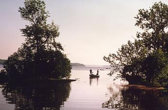 A photograph of the Potomac river. There are two trees on either side of the image. There looks to be two men on a dock in the distance. The water is flat. 