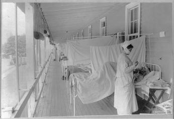 A black and white photograph of a nurse caring for a line of men stricken with the Spanish Flu at Walter Reed Hospital. They are on a covered porch, the Nurse wears  1910s personal protective equipment. 