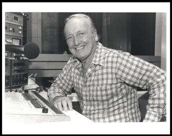 An older Grant sits at a desk in a radio station booth. He has an elbow on the table and gives a big smile to the camera.