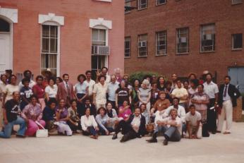 A photograph of a large group of people sitting and standing outside of a brick building