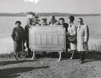 A group of 7 people standing around a sign. Three are women. Turkey Tayac can be seen with a Native American headdress. In the background can be seen the Potomac River.