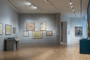A photo of a gallery space at the GW Museum. Pieces from Mr. Small’s collection adorn the pale blue walls. In right background, a portrait of Mr. Small hangs proudly.
