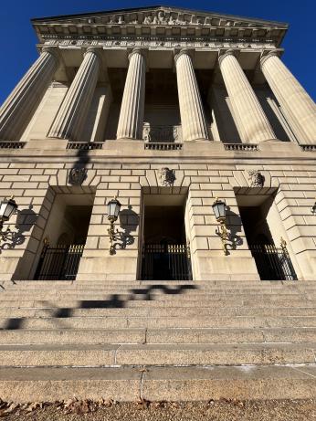A photo of the outside of the Andrew W. Mellon Auditorium building today. It is made of a sand-colored stone and in a Greek revival style. There are about 10 stairs leading up to three large gated entrances. Above the entrance are several columns and an ornately carved pointed roof.