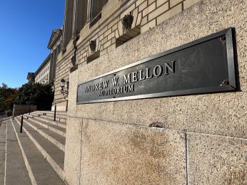 A closeup photo of a metal sign that reads “Andrew W. Mellon Auditorium.” The sign is mounted on a sand-colored stone wall. Past the sign, stairs and the front of the building are visible. 
