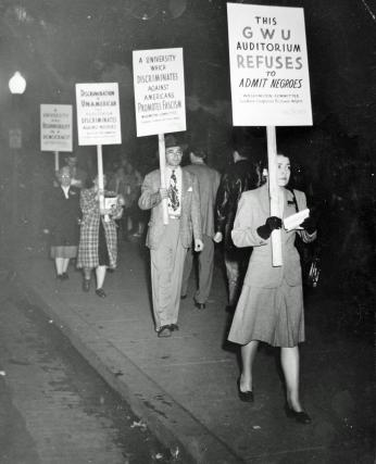 A photograph of people protesting. They are both men and women and they wear clothes from the 1940s. They hold signs that say things such as “a university that discriminates against Americans promotes fascism.”