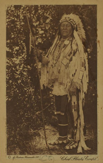 Photograph of Chief Plenty Coups in his war bonnet and holding a coup stick.