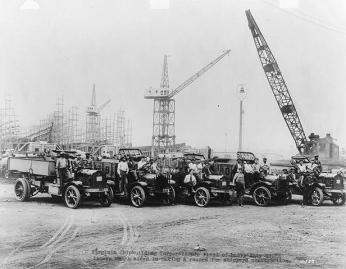 Five early 20th century trucks in the foreground with many men sitting in the front seats and a shipyard with cranes in the background 