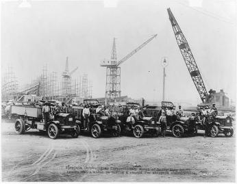 Five early 20th century trucks in the foreground with many men sitting in the front seats and a shipyard with cranes in the background 