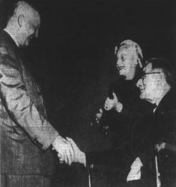 A grainy black-and-white photo. Eisenhower stands and shakes hands with Deffner who is in his wheelchair. Behind Deffner, his wife stands holding a trophy. All three are smiling.