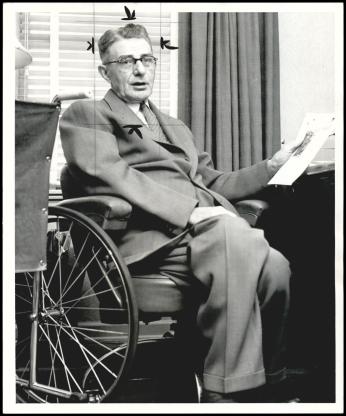 Hugo Deffner on April 4, 1957 (Jim Lucas, Oklahoma Historical Society) A black-and-white photo. Hugo Deffner, a white man in his 60s, sits in his wheelchair at a desk. He is holding a piece of paper in one hand and wearing a suit. He is looking at the camera.