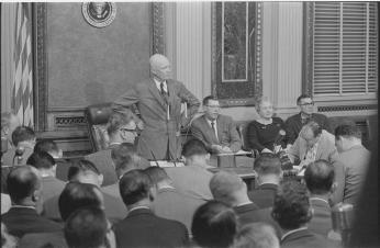 President Eisenhower stands at a table at the White House during a news conference, surrounded by the heads of mostly male reporters.