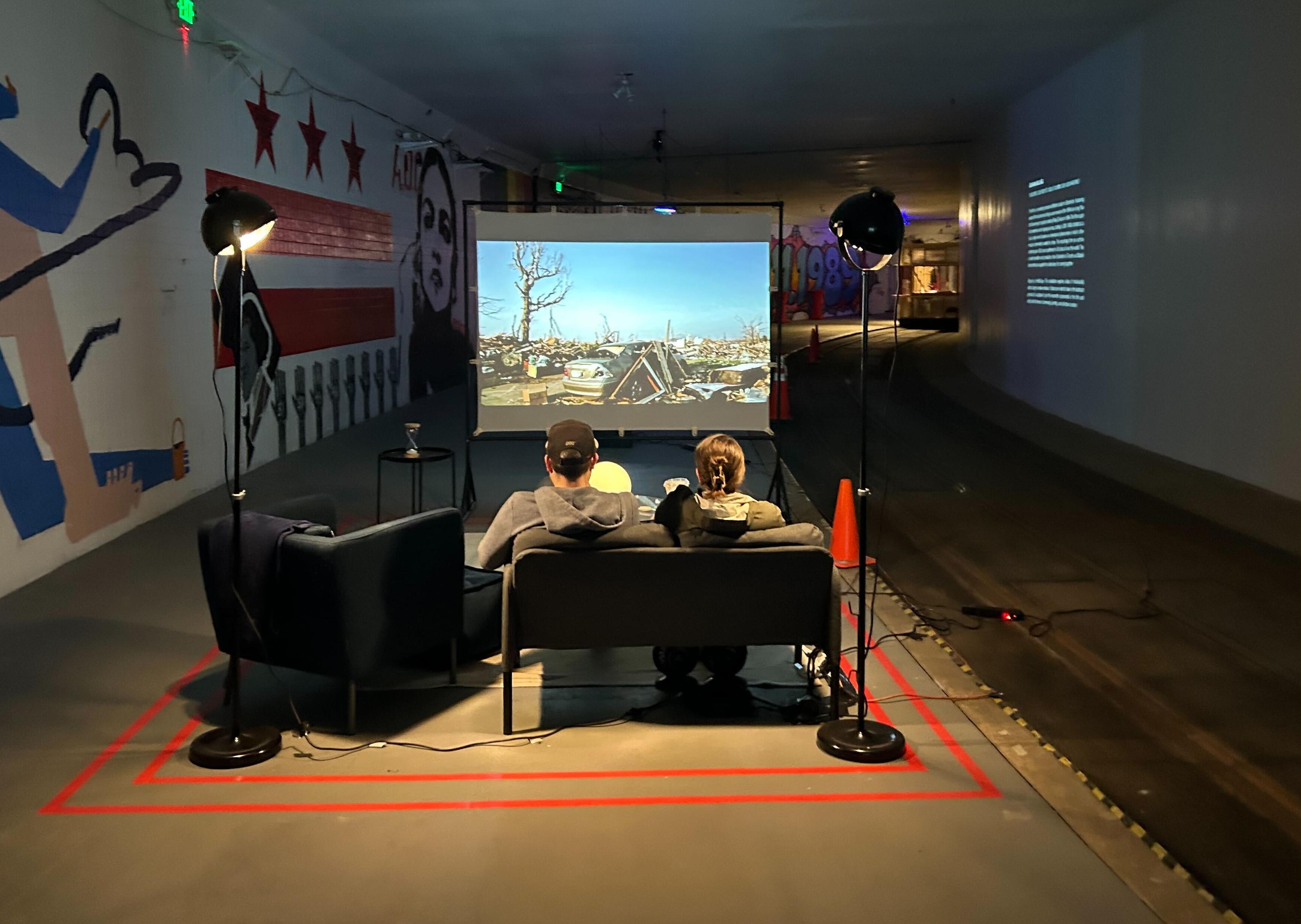 A couple sit on a couch, watching a film projected on a screen before them. Unusual about the image, is that the couch and the rest of this living room like set-up, are actualy stationed in a tunnel beneath Dupont Circle. Old streetcar tracks are to their right, while graffiti murals adorn the brick walls to their left.